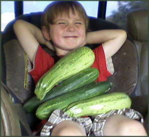 a grinning child sits with 2 zucchinis and 2 cucumbers spread across his chest, nearly covering him