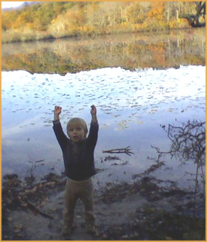 a small child singing and with arms outstretched above his head with a still pond in the background reflecing fall colors